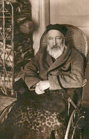 Jonas Jablonskis at home (~1920–1925) (Maironis Lithuanian Literature Museum, No. MLLM F1 17394). A man approximately 60–65 years old with a beard, wearing a robe and a black cap, sitting in a wheelchair, covered with a patterned blanket, with his arms crossed over it. Next to him is a shelving unit filled with documents and papers.