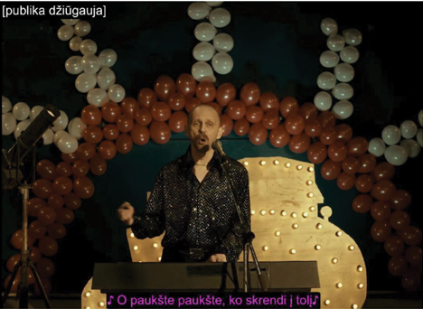 In a festive setting, a man with a short beard stands at a keyboard, wearing a sparkly black shirt. Behind him is a large decorative arrangement of red and white balloons shaped into arches and a lit stage prop. A microphone is positioned in front of him, and he appears to be singing. Subtitles at the bottom of the image show the Lithuanian lyrics “O paukšte paukšte, ko skrendi į toli” (Oh bird, oh bird, why are you flying off into the distance), while the caption at the top reads “publika džiūgauja” (the audience rejoices).