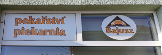 Zdjęcie przedstawia fragment witryny sklepowej. W prawej części jest nazwisko/nazwa sklepu „Bajusz” i logo rogalika (pieczywo). Wlewo są dwa napisy – u góry czeski „pekařství”, pod nim polski „piekarnia”. Graficznie są wszystkie napisy tożsame – jednakowej wielkości i pomarańczowego koloru.
