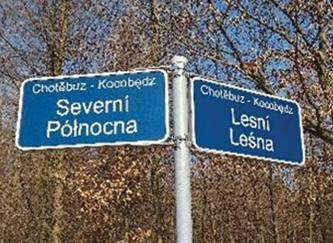 [Dwa zdjęcia obok siebie. Na pierwszym zdjęciu znajduje się fragment elewacji domu, na którym jest dwujęzyczna tablica z nazwą ulicy – Alšova / Aleša. Tabliczka to metalowy prostokąt, na niebieskim tle widinieje biała czcionka. Drugie zdjęcie przedstawia dwie tablice z nazwmi ulic zainstalowanymi na jednym metalowym słupku w otoczeniu krzewów i drzew. Na tablicy wskazującej wlewo jest mniejszy napis „Chotěbuz – Kocobędz” u góry, natomiast w środku większą czcionką napis „Severní / Pólnocna”.  Na tablicy wskazującej wprawo jest mniejszy napis „Chotěbuz – Kocobędz” u góry, natomiast w środku większą czcionką napis „Lesní / Leśna”. Tabliczki to metalowe prostokąty, na niebieskim tle widinieje biała czcionka.]
