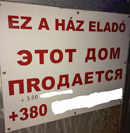Фото 5. На белой табличке крупными красными буквами написан текст на венгерском и на русском: “EZ A HÁZ ELADÓ” и ниже «ЭТОТ ДОМ ПРОДАЕТСЯ». В слове «ПРОДАЕТСЯ» используется латинская буква “R” вместо кириллической «Р», что является ошибкой. Ниже указаны два украинских номера, начинающиеся с кода «+380» (последующие цифры скрыты автором этой статьи для сохранения конфиденциальности).