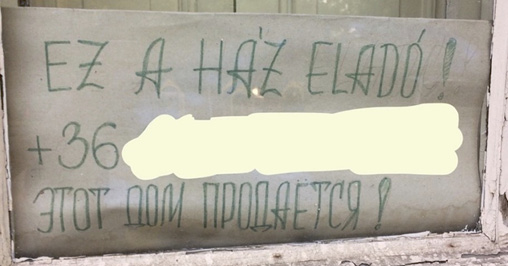 Фото 4. На окне частного дома размещён лист бумаги с рукописным объявлением зелёными буквами. Верхняя строка этого объявления написана на венгерском: “EZ A HÁZ ELADÓ!”. На второй строке указан венгерский номер телефона, начинающийся с кода «+36» (последующие цифры скрыты автором этой статьи для сохранения конфиденциальности). Внизу по-русски написано: «ЭТОТ ДОМ ПРОДАЁТСЯ!». В этом объявлении нет грамматических ошибок.