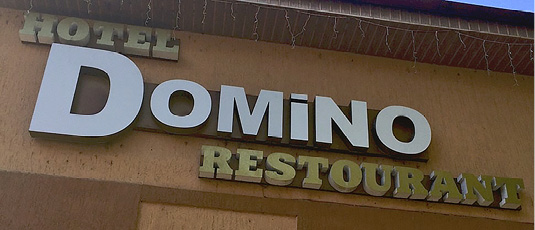 Фото 9. На стене дома коричневого цвета установлена вывеска гостиничного комплекса с рестораном на английском языке. Слева вверху золотыми буквами написано слово “HOTEL”. По центру заметно большими по шрифту белыми буквами указано название заведения – “Domino”. В правой нижней части фотографии присутствует слово “RESTAURANT”, написанное с ошибкой (вместо первой буквы “A” здесь используется “O”). Слово “RESTAURANT” оформлено в таком же стиле и таким же шрифтом, что и “HOTEL”.