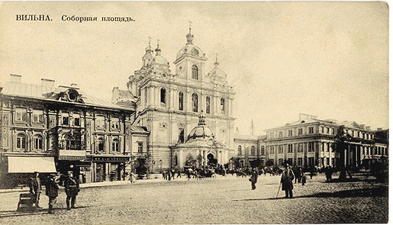 A historic sepia-toned street scene shows a large ornate church with a dome and towers in the center, flanked by multi-story buildings, with people walking and standing on a cobblestone square.

Description generated by AI
