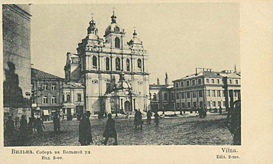 A historic city square with a large ornate baroque-style church in the center, surrounded by several people walking and standing on a cobblestone street.

Description generated by AI