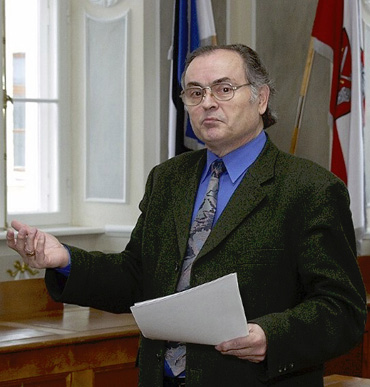 A person wearing glasses, a dark blazer, blue shirt, and patterned tie is holding papers and gesturing with one hand in a formal room with flags and large windows.

Description generated by AI