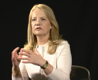 A person with long hair wearing a light-colored top is speaking and gesturing with their hands against a dark background.

Description generated by AI